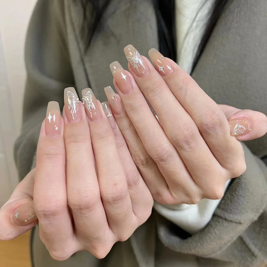 A pair of hands showing long coffin-shaped nails in a glossy nude pink. Each nail has delicate silver butterfly and star decals. The wide, flat tips and tapered sides create a sleek, elongated look.